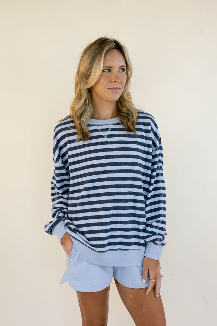 A woman modeling a navy and light blue horizontally striped long-sleeve sweatshirt with matching light blue shorts, standing against a plain beige background.
