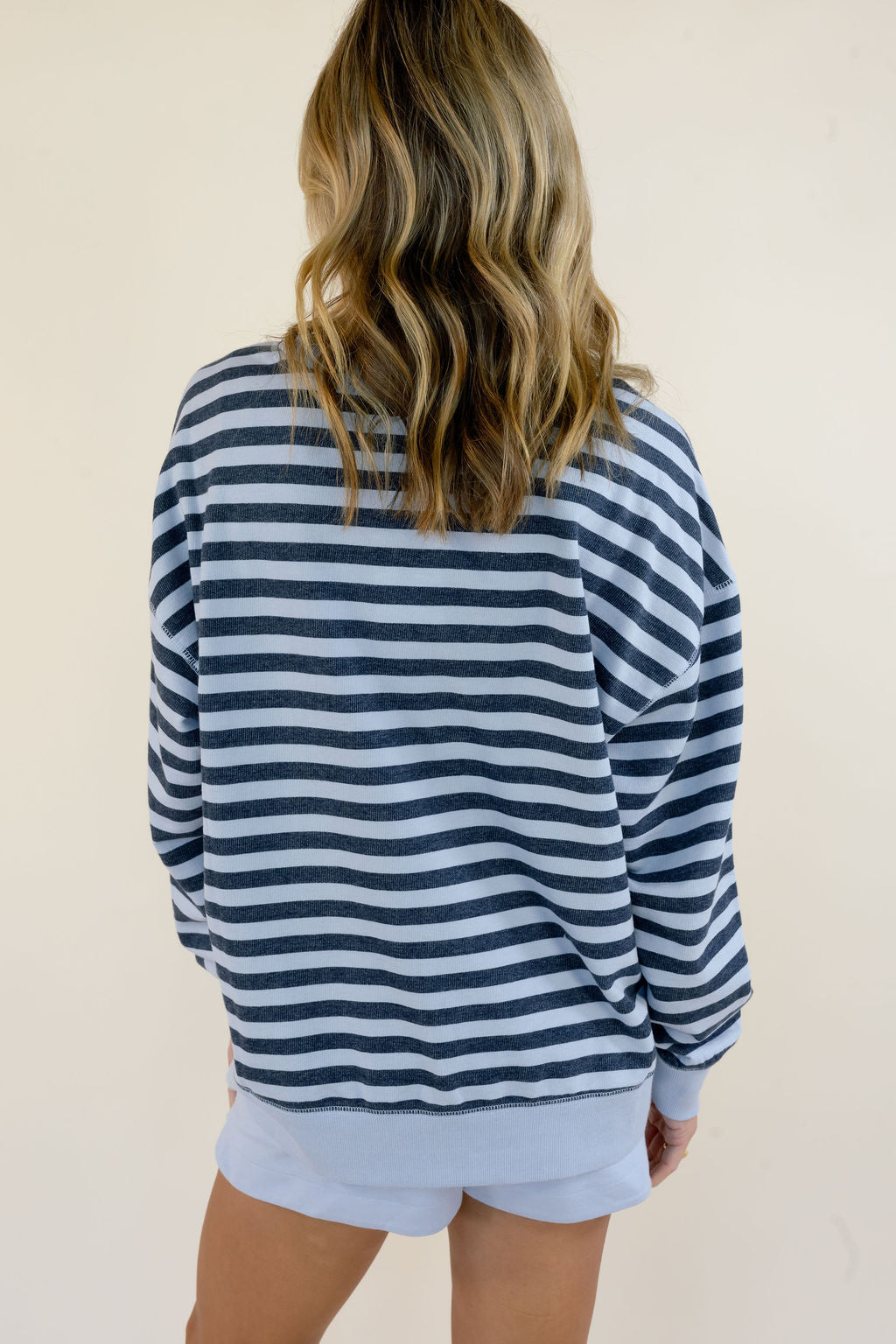 A woman viewed from behind wearing a navy blue and light blue horizontally striped long-sleeve sweatshirt and matching light blue shorts against a plain beige background.