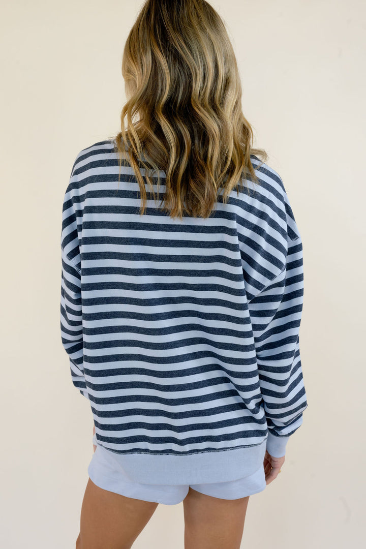 A woman viewed from behind wearing a navy blue and light blue horizontally striped long-sleeve sweatshirt and matching light blue shorts against a plain beige background.