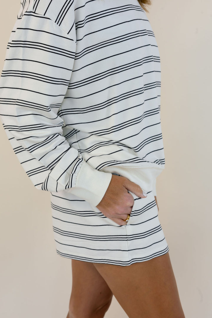 A close-up of a person wearing a white long-sleeve sweatshirt and shorts set with thin black horizontal stripes, standing against a plain beige background.