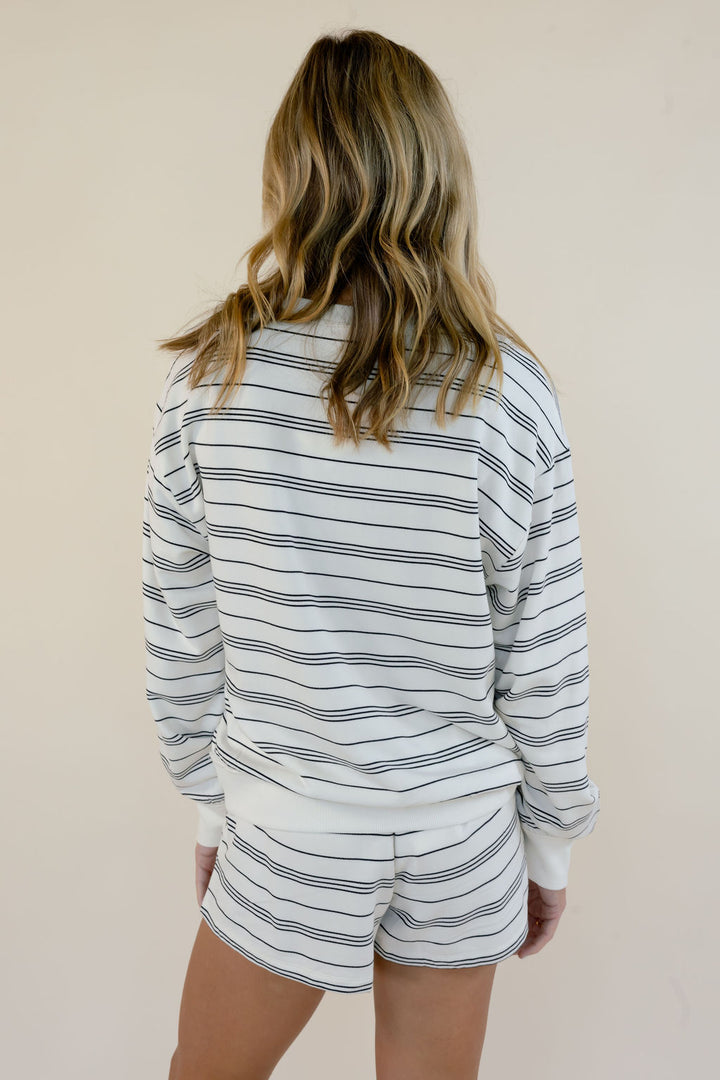 A woman viewed from behind wearing a matching white lounge set with thin black horizontal stripes, consisting of a long-sleeve sweatshirt and shorts, against a plain beige background.