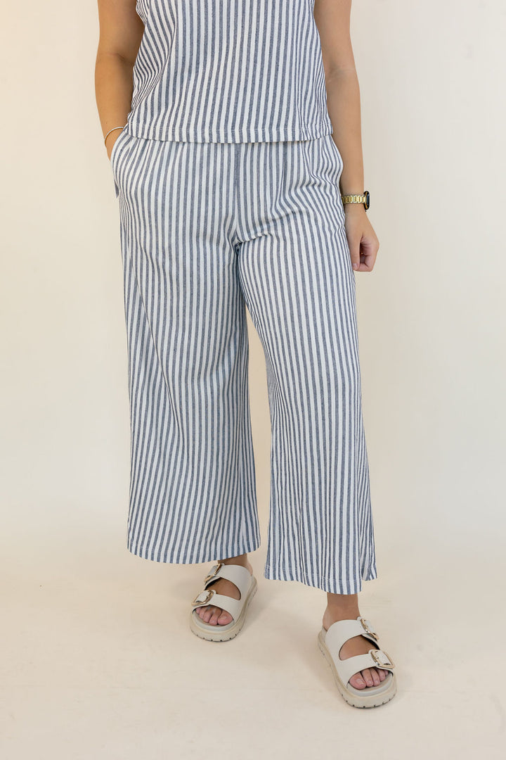 A person wearing a matching sleeveless top and wide-leg cropped pants set in a blue and white vertical stripe fabric, paired with white slide sandals, photographed against a plain off-white studio background.