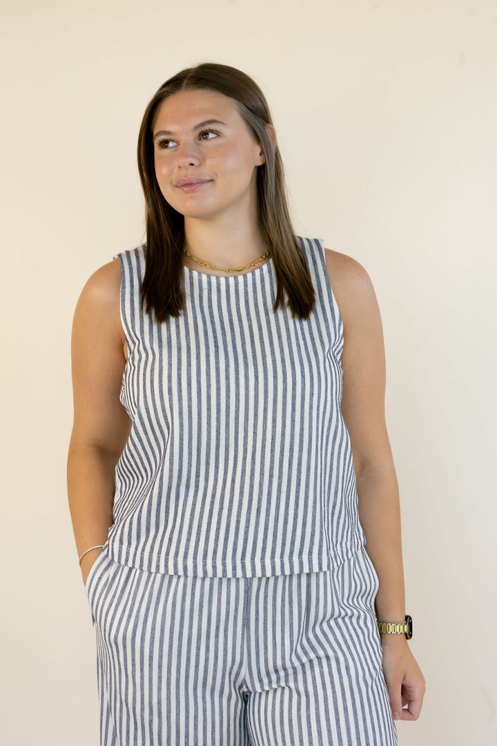 A woman wearing a sleeveless top and matching striped pants in a blue and white vertical stripe pattern, standing against a plain off-white wall.