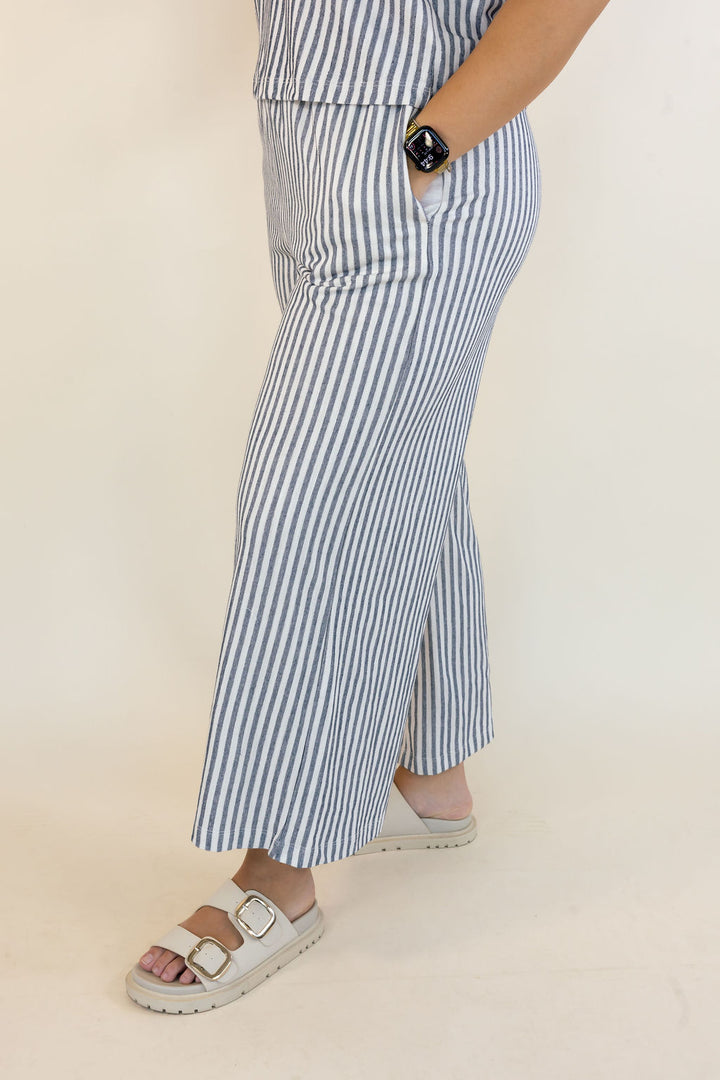 A person wearing blue and white vertically striped wide-leg cropped pants with side pockets, paired with white slide sandals, shown from the waist down against a plain cream background.