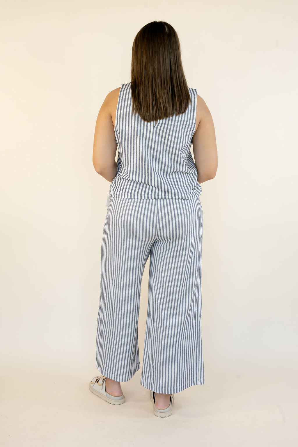 A woman viewed from behind wearing a sleeveless blue and white vertically striped matching set with wide leg pants, standing against a plain off-white studio background.