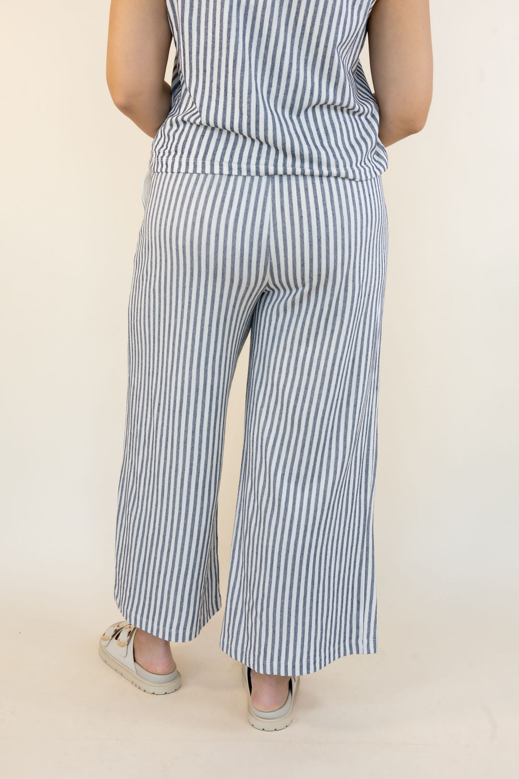 A person shown from the back wearing a matching sleeveless top and wide-leg pants set in a blue and white vertical stripe pattern, paired with beige sandals, against a plain cream background.
