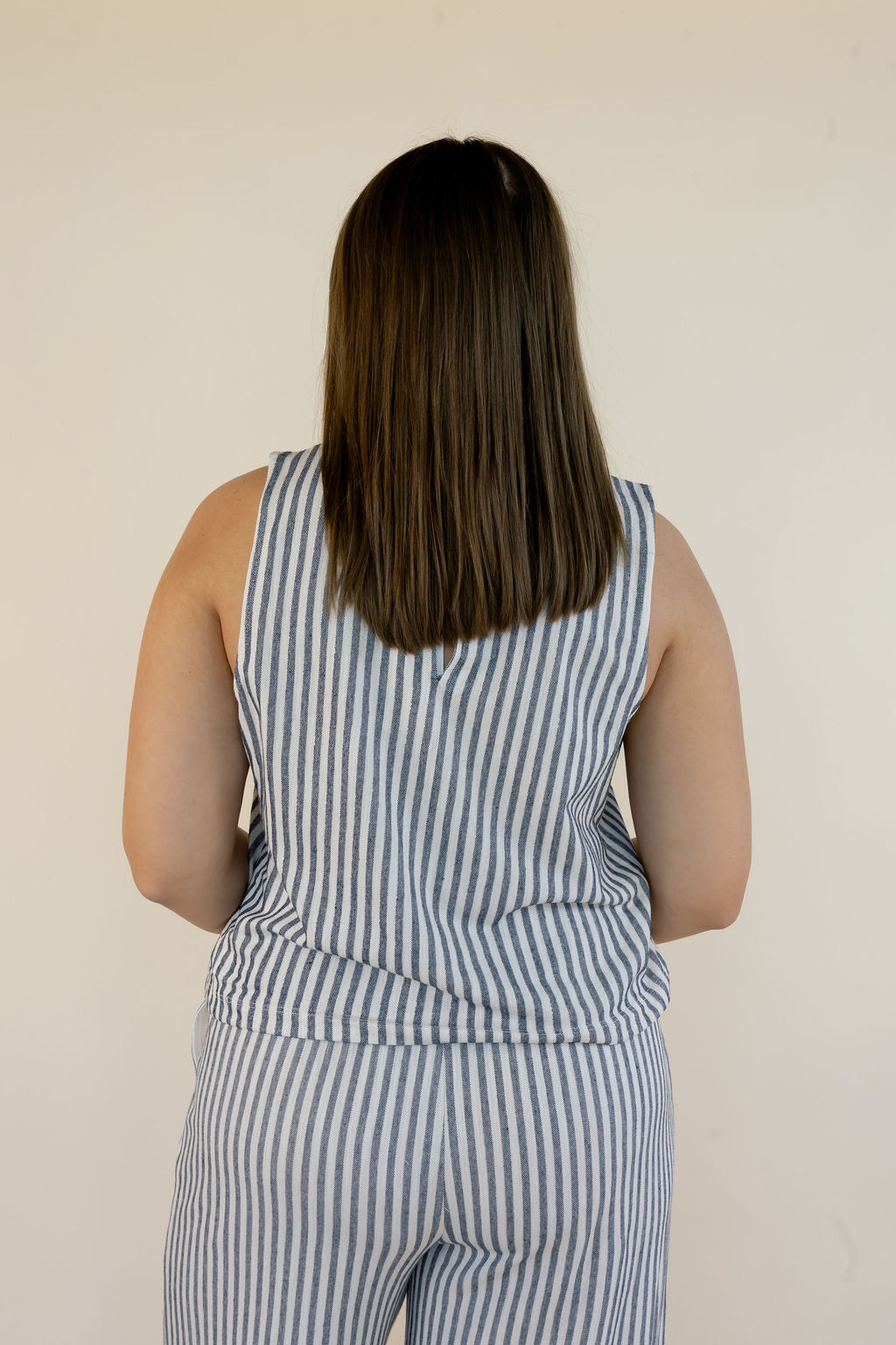 A woman viewed from behind wearing a sleeveless blue and white vertically striped matching set against a plain off-white wall.