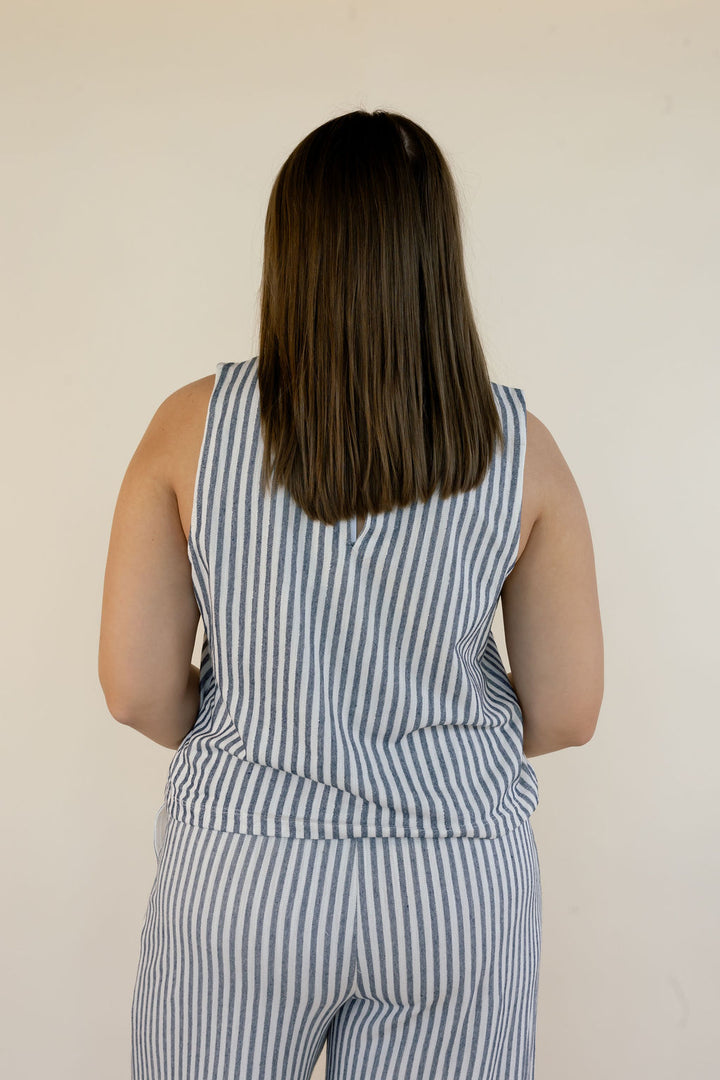 A woman viewed from behind wearing a sleeveless blue and white vertically striped matching set against a plain off-white wall.