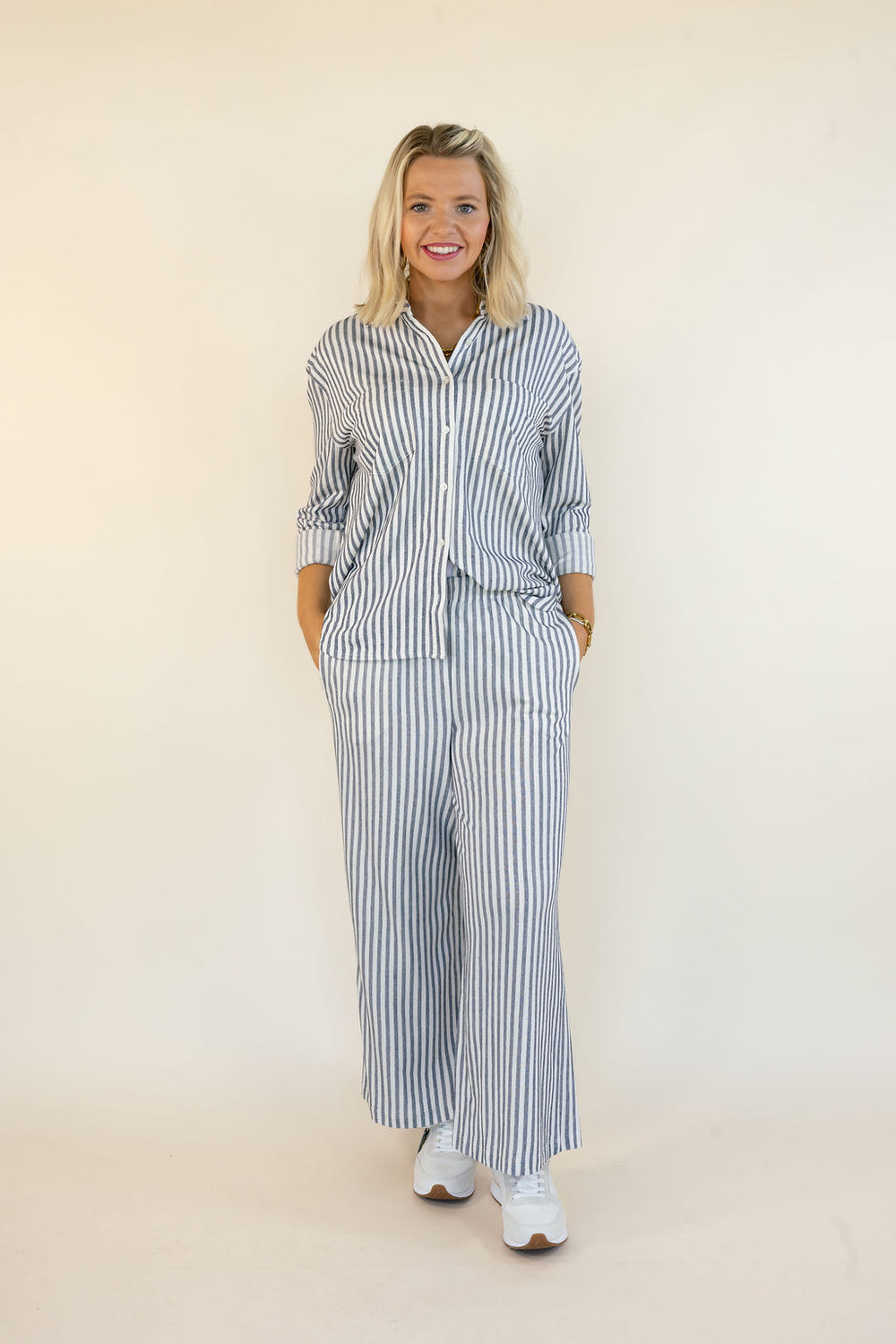 A woman modeling a matching two-piece set with vertical blue and white stripes, featuring a long-sleeve button-up shirt and wide-leg pants, standing against a plain cream background.