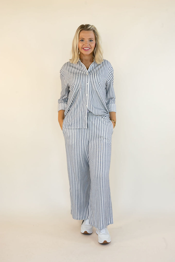 A woman modeling a matching two-piece set with vertical blue and white stripes, featuring a long-sleeve button-up shirt and wide-leg pants, standing against a plain cream background.
