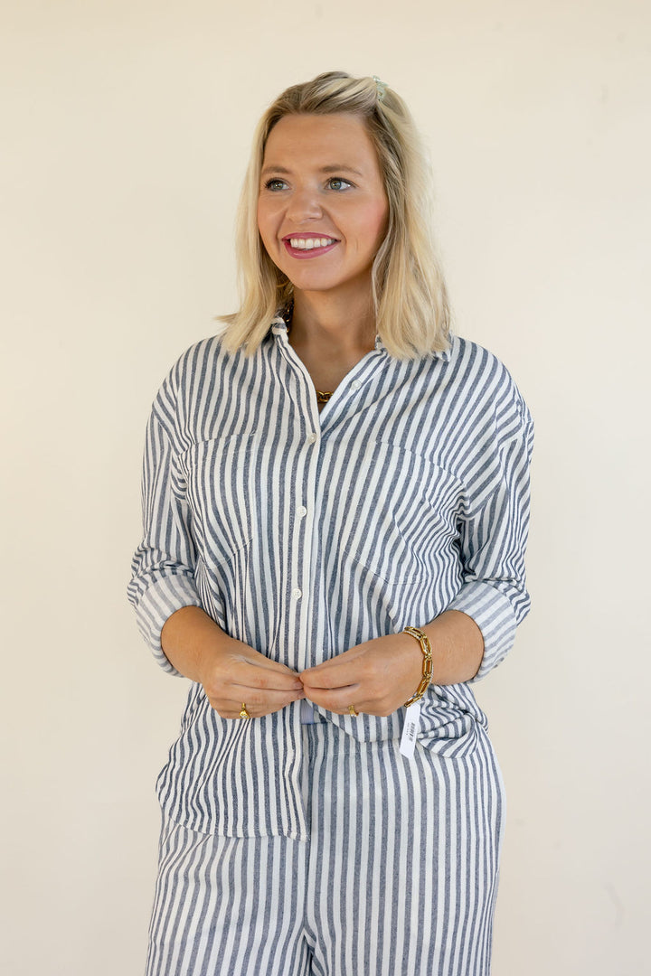 A smiling blonde woman wearing a blue and white vertically striped button-up shirt and matching striped pants, standing against a plain off-white background.