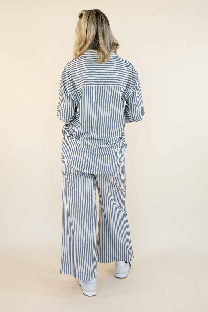A woman viewed from behind wearing a matching blue and white vertically striped long-sleeve top and wide-leg pants set, paired with white sneakers, against a plain beige studio background.