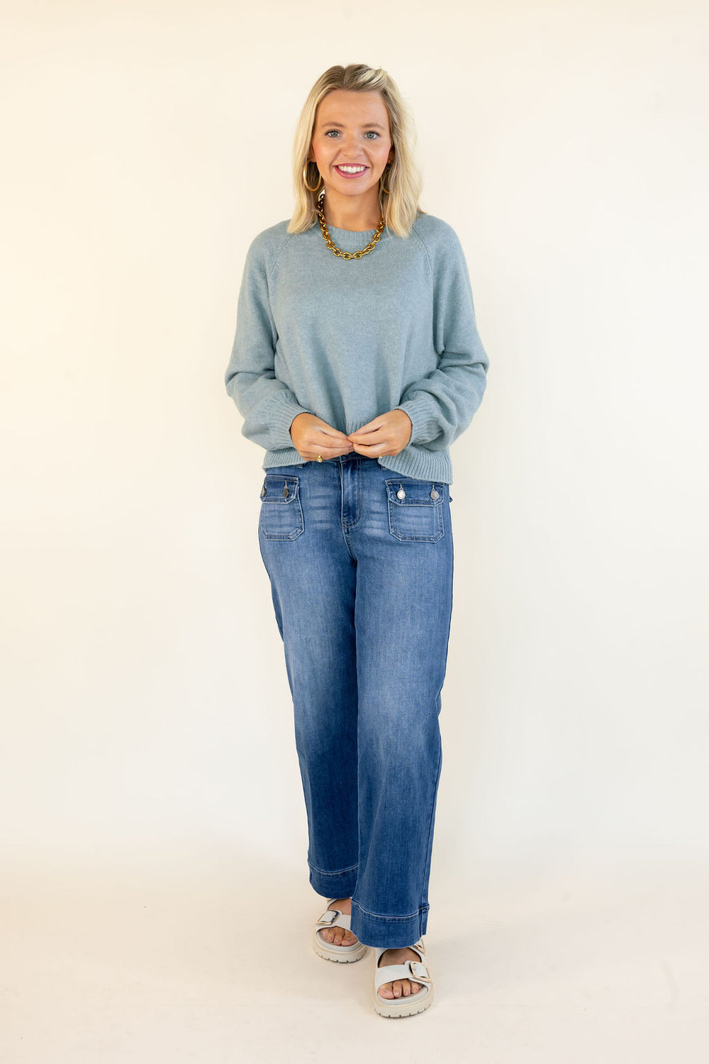 A woman modeling a blue-green crewneck sweater and blue jeans against a plain white studio background.