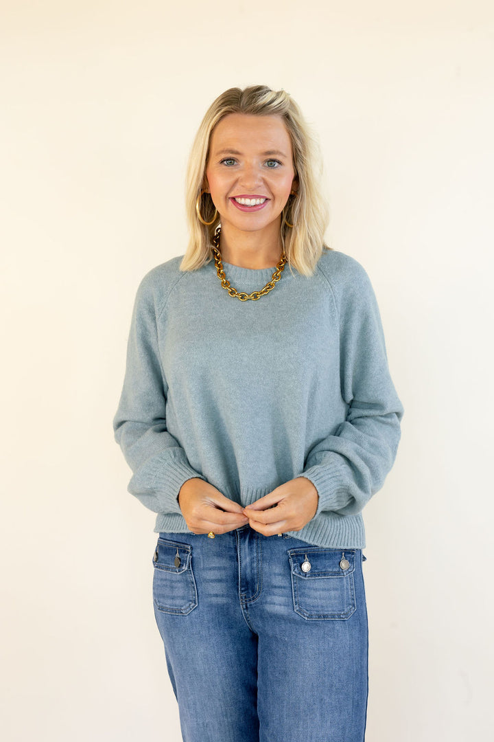 A smiling blonde woman wearing a light blue-green knit sweater and blue jeans, standing against a plain off-white background.
