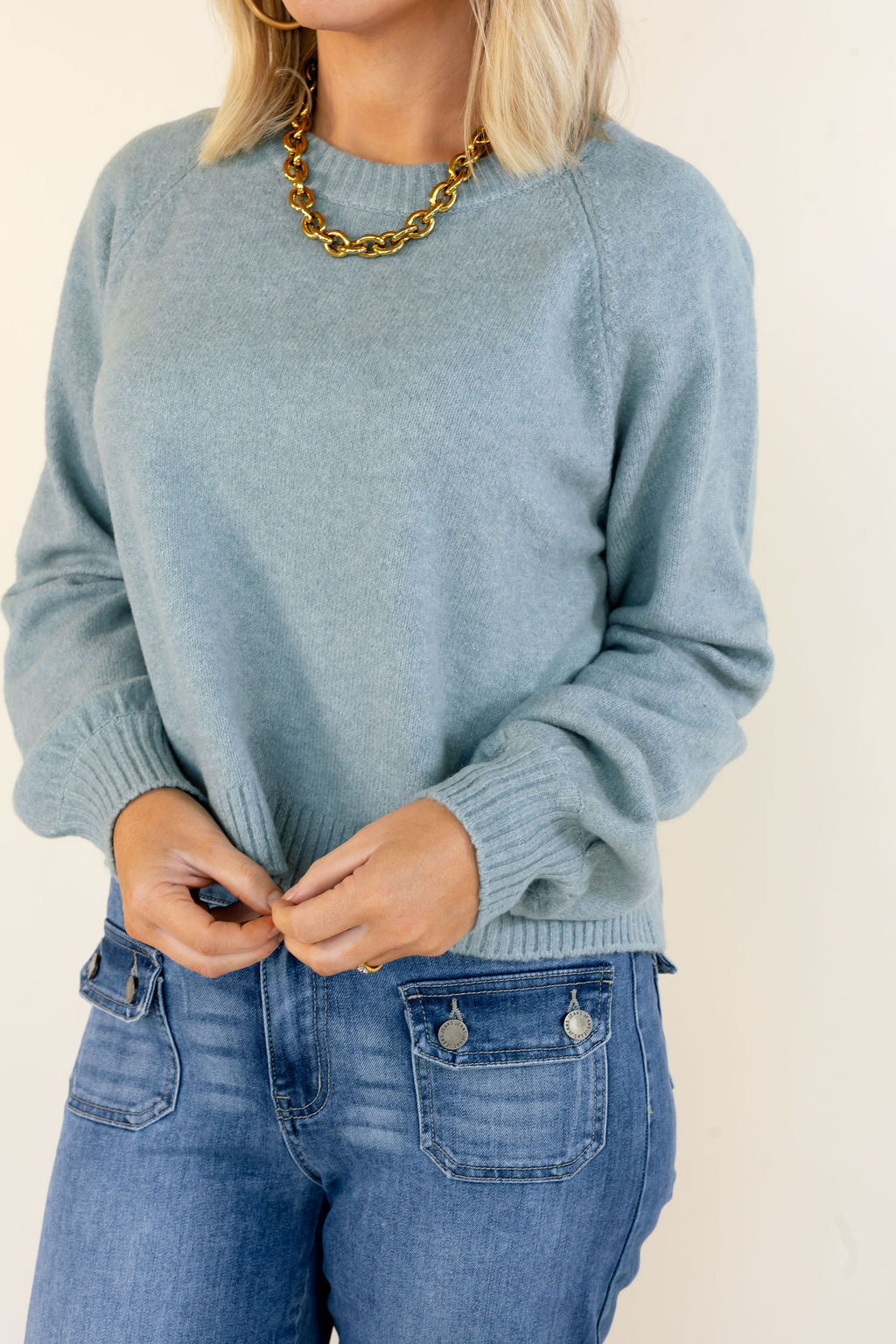 A woman wearing a light blue-green knit crewneck sweater and blue jeans, accessorized with a chunky gold chain necklace, against a plain off-white background.