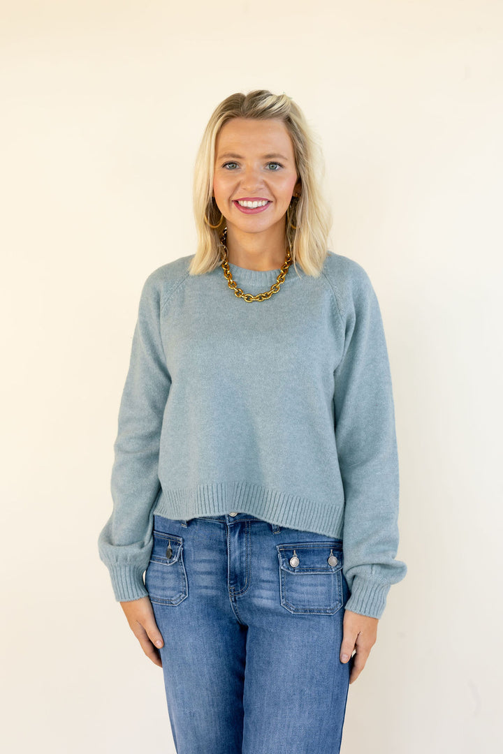 A smiling blonde woman wearing a light blue-green knit crewneck sweater and blue jeans, standing against a plain off-white background.