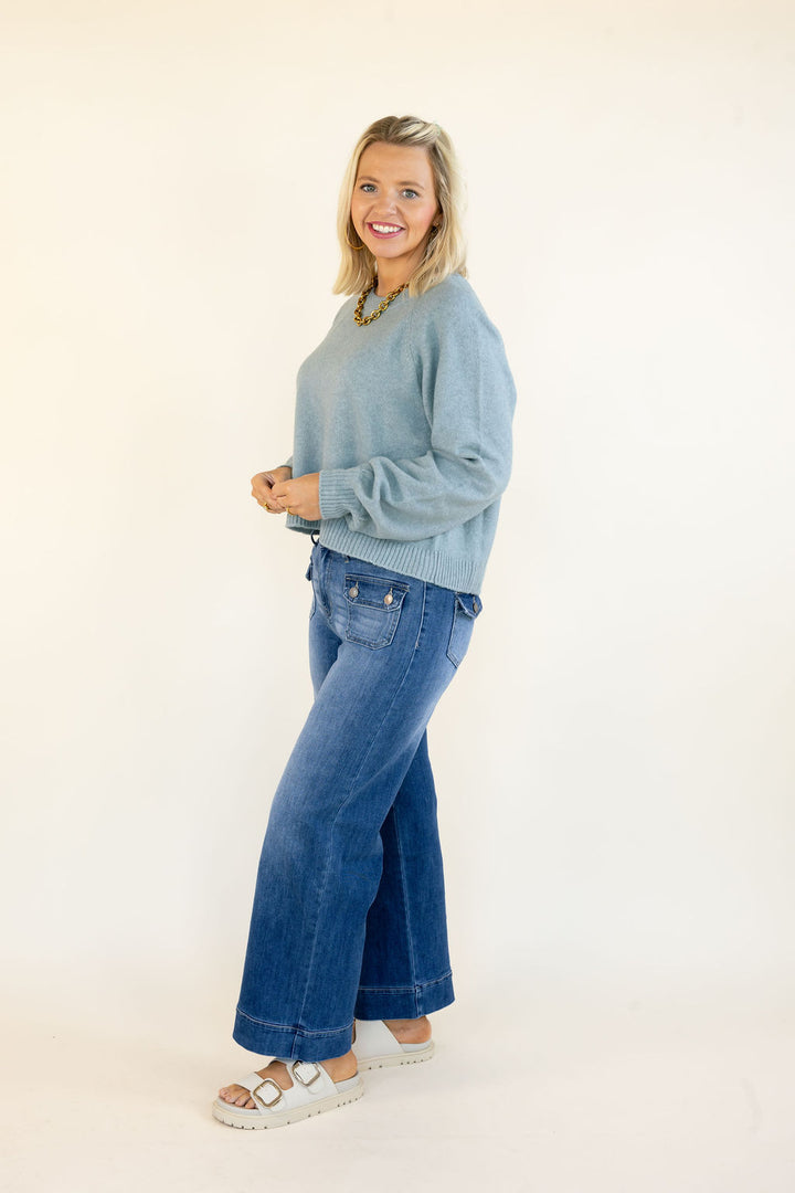 A woman modeling a light blue-green knit sweater and wide-leg blue jeans with white sandals, standing against a plain off-white studio background.