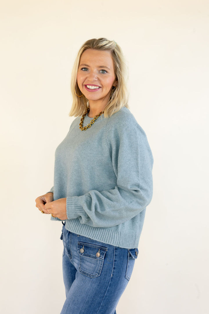 A smiling blonde woman wearing a light blue-green knit sweater and blue jeans, standing against a plain off-white background.