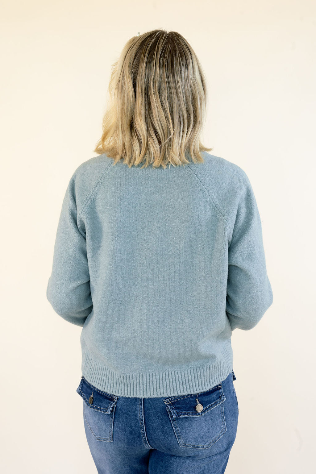 A woman viewed from behind wearing a light blue-green knit sweater and blue jeans against a plain off-white background.