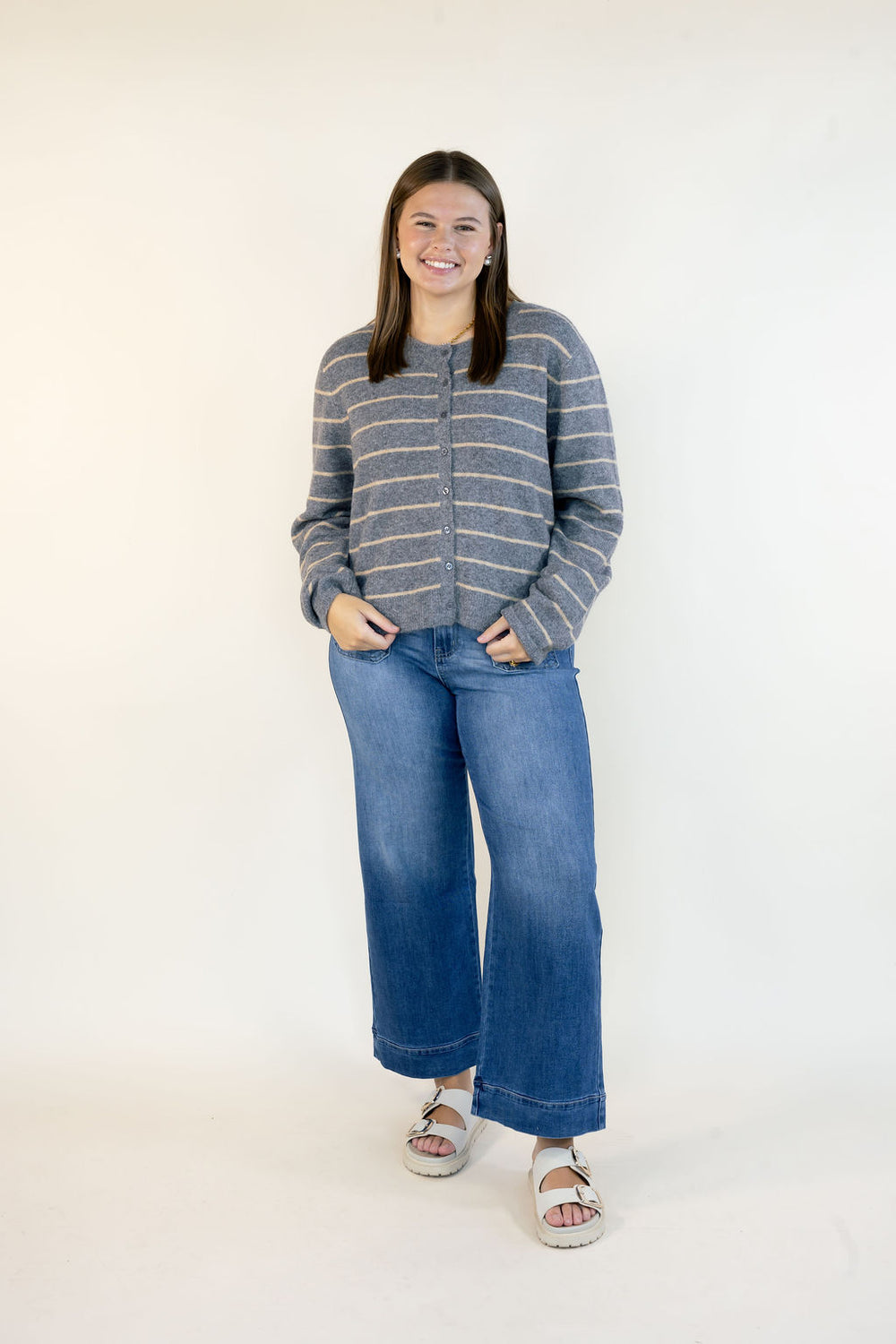 A woman modeling a gray and beige striped knit cardigan sweater with wide-leg blue jeans and white slide sandals, standing against a plain white studio background.