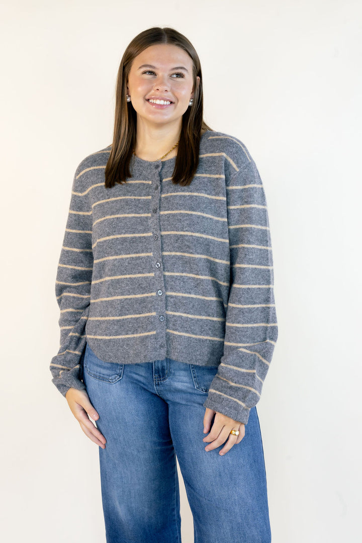 A woman wearing a gray and beige striped knit cardigan sweater with blue jeans, standing against a plain white background.