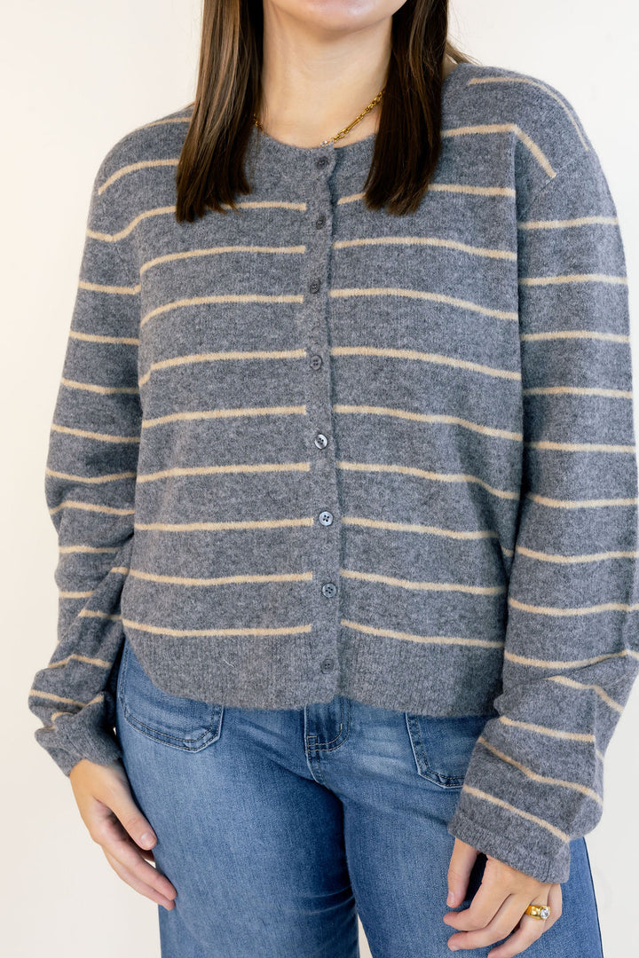 A woman wearing a gray knit cardigan with thin beige horizontal stripes and blue jeans, shown from the neck down against a plain light background.