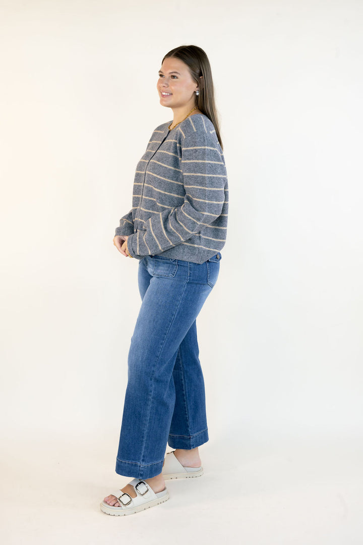 A woman modeling a gray and beige striped knit sweater with blue wide-leg jeans and white slide sandals, standing against a plain white studio background.