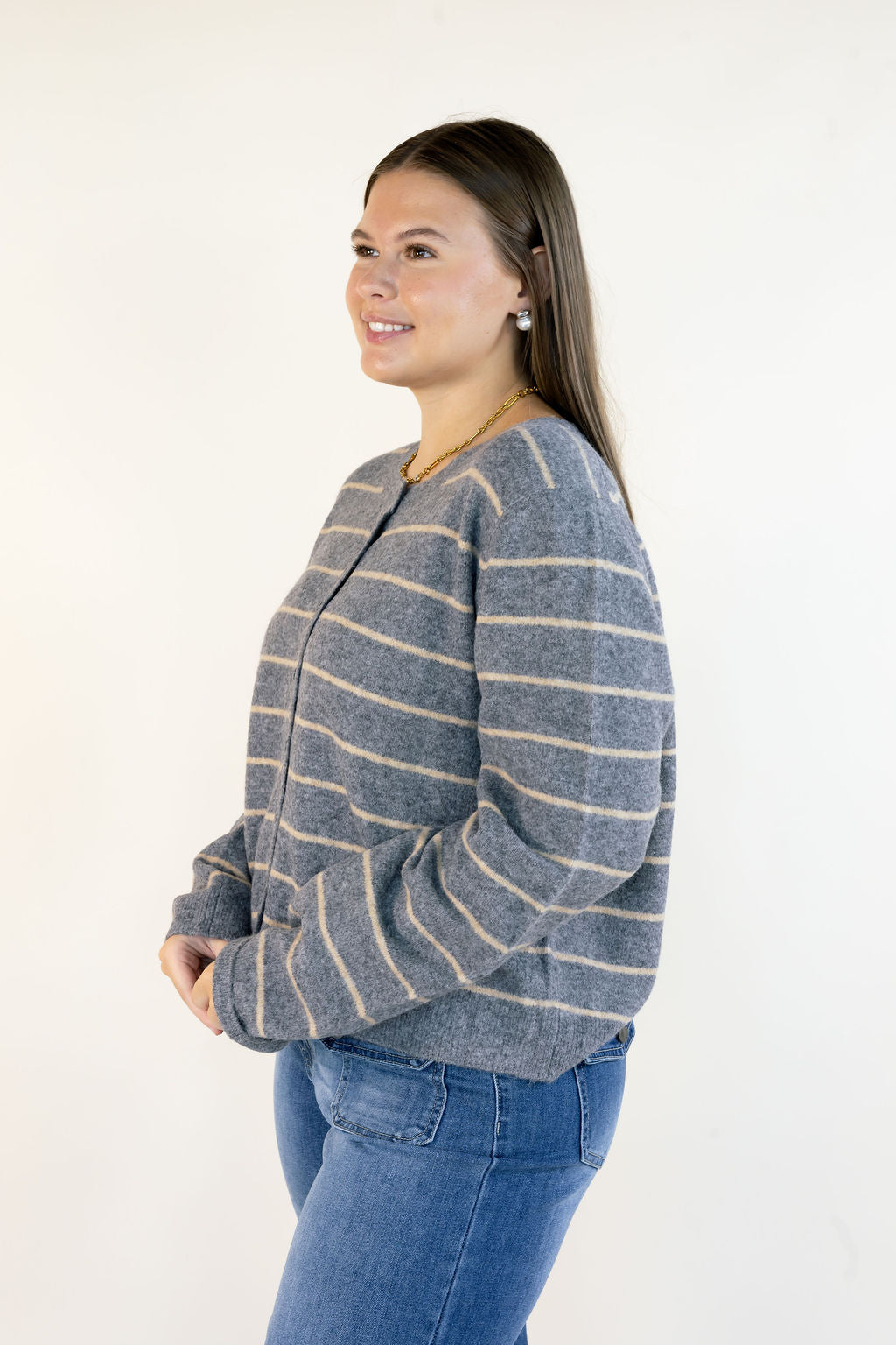 A woman wearing a gray knit cardigan with thin beige horizontal stripes and blue jeans, standing against a plain white studio background.