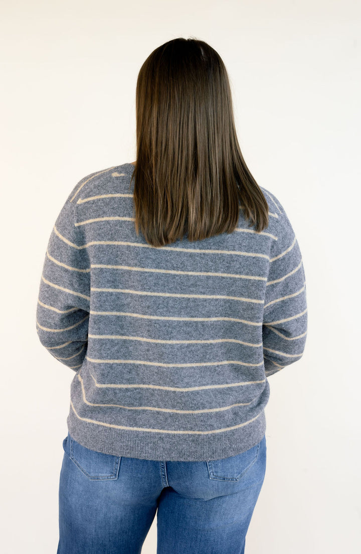 A woman viewed from behind wearing a gray knit sweater with thin beige horizontal stripes and blue jeans, against a plain off-white background.