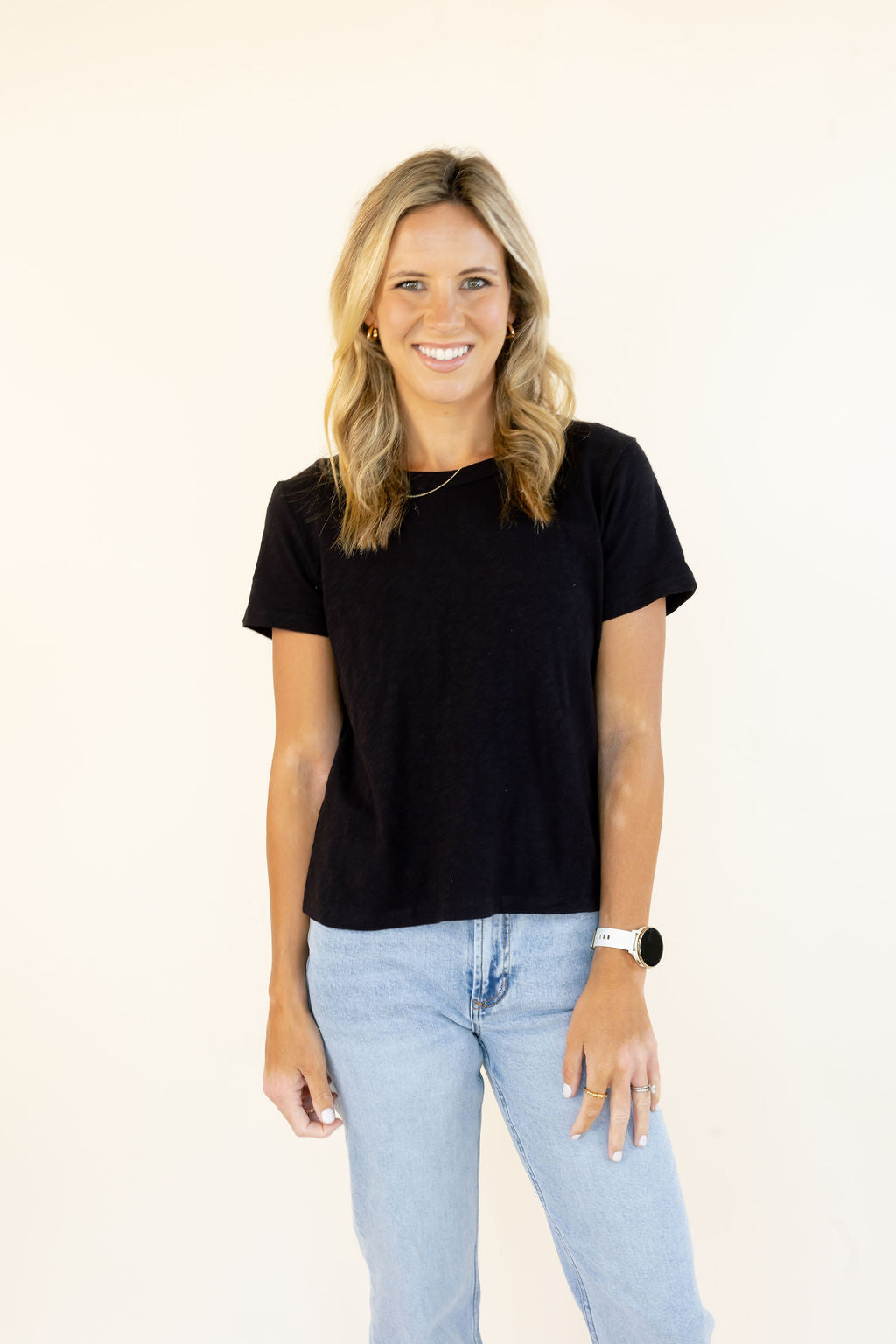 A smiling blonde woman wearing a black short-sleeve crewneck t-shirt and light blue jeans, standing against a plain off-white background.