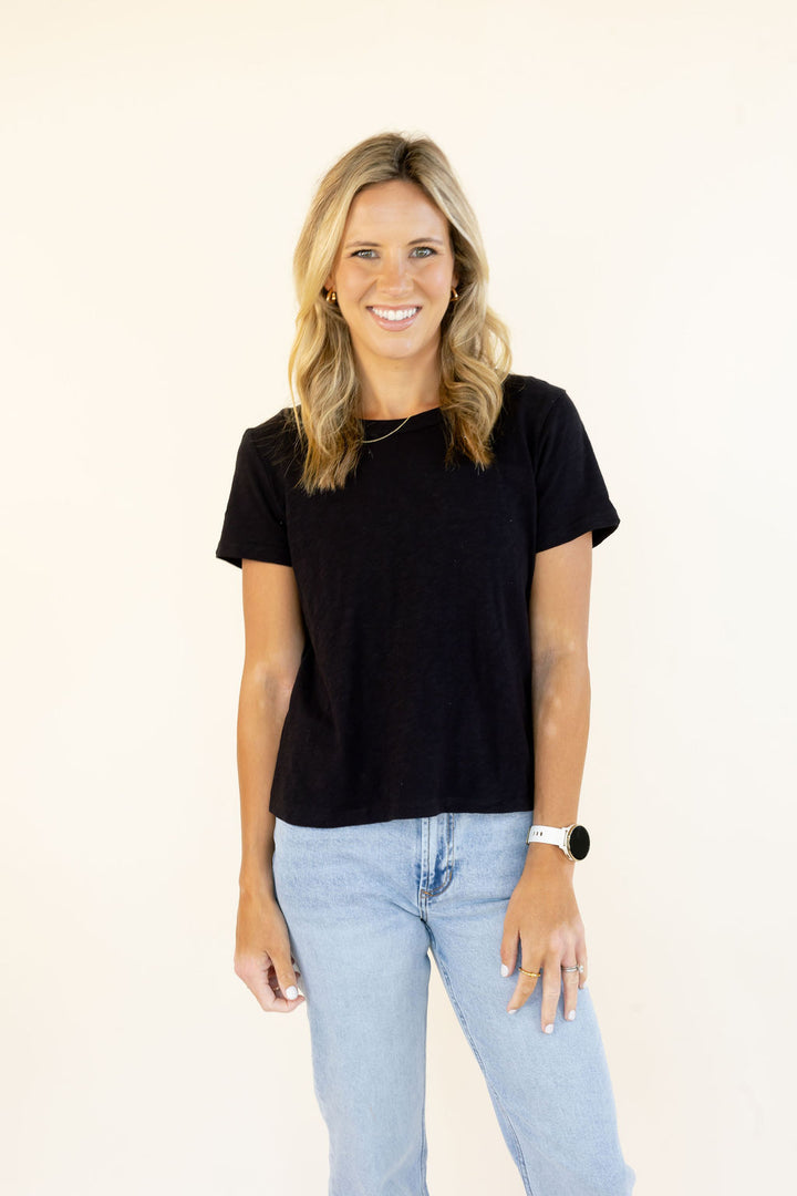 A smiling blonde woman wearing a black short-sleeve crewneck t-shirt and light blue jeans, standing against a plain off-white background.