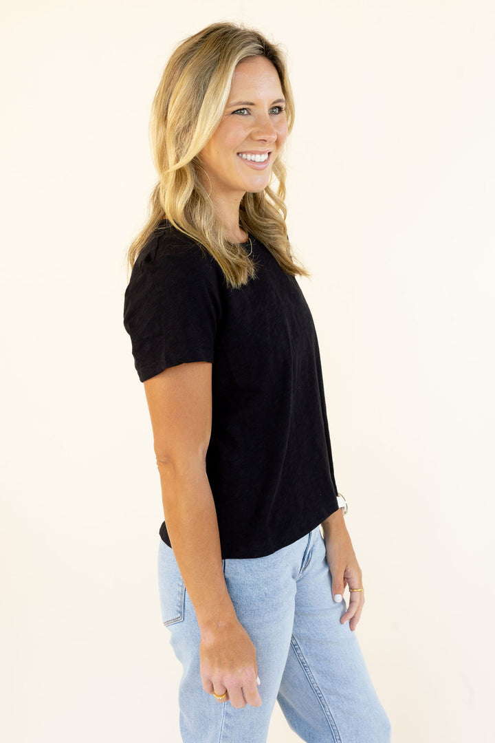 A woman with blonde hair wearing a black short-sleeve crewneck t-shirt and light blue jeans, standing against a plain off-white background.