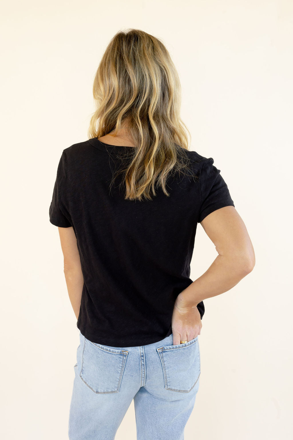 A woman with blonde-highlighted hair is shown from behind wearing a black short-sleeve t-shirt and light-wash blue jeans against a plain off-white background.