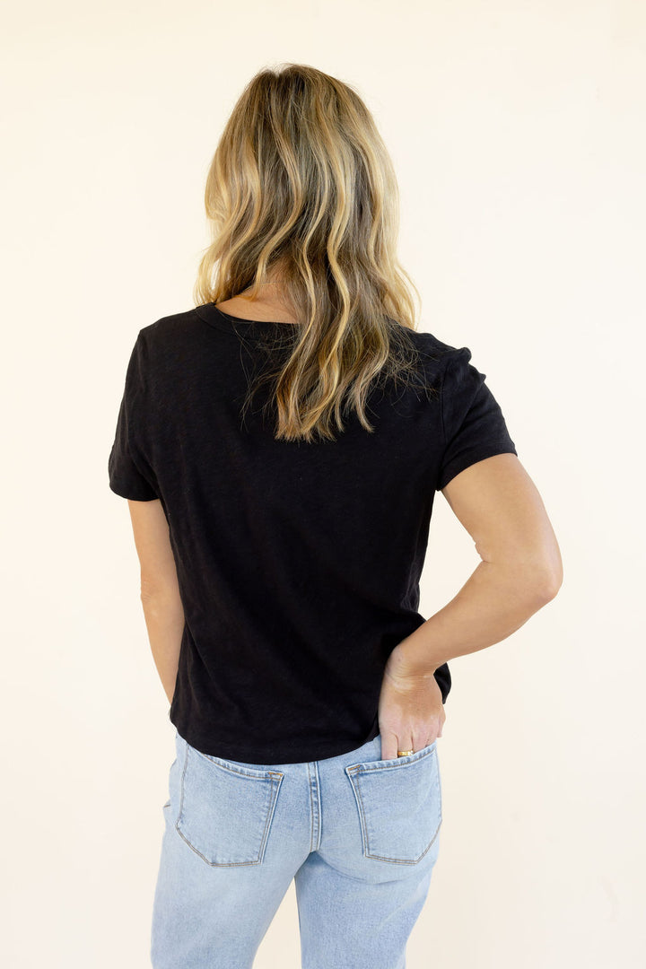 A woman with blonde-highlighted hair is shown from behind wearing a black short-sleeve t-shirt and light-wash blue jeans against a plain off-white background.