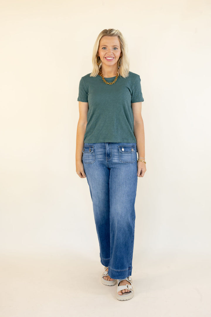 A woman modeling a dark green short-sleeve t-shirt and wide-leg blue jeans, standing against a plain off-white studio background.