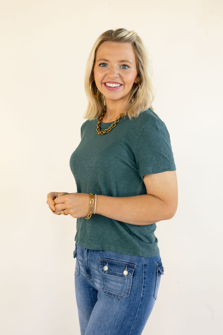 A smiling blonde woman wearing a dark green short-sleeve t-shirt and blue jeans, standing against a plain off-white background.