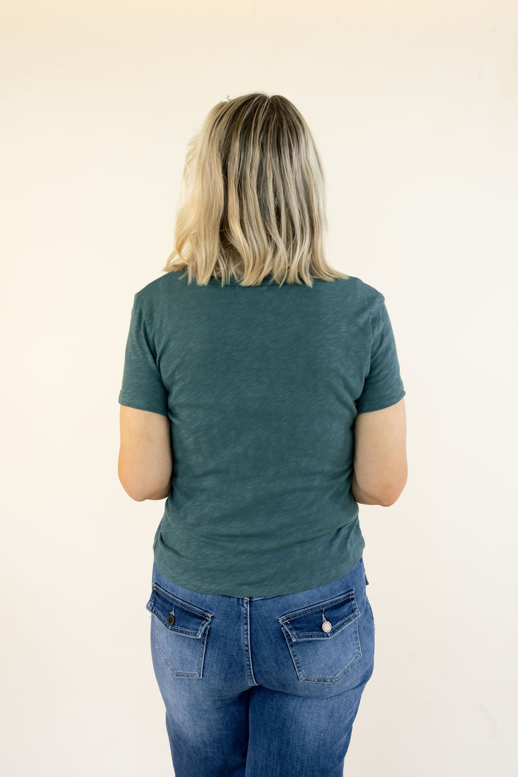 A woman viewed from behind wearing a dark teal-green short-sleeve t-shirt and blue jeans, standing against a plain off-white background.