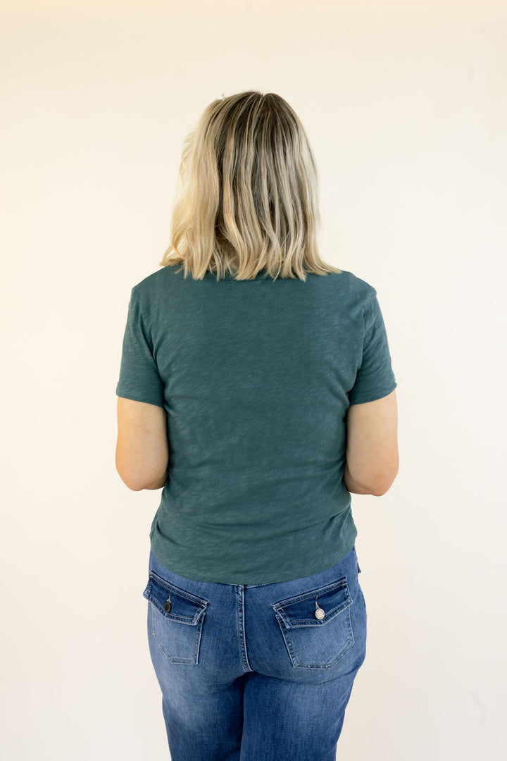 A woman viewed from behind wearing a dark teal-green short-sleeve t-shirt and blue jeans, standing against a plain off-white background.