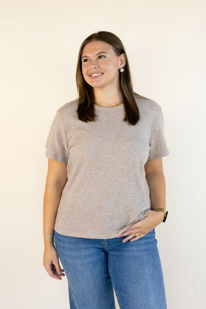 A woman modeling a short-sleeve crewneck t-shirt in a light heather ash color, paired with blue jeans, standing against a plain off-white background.