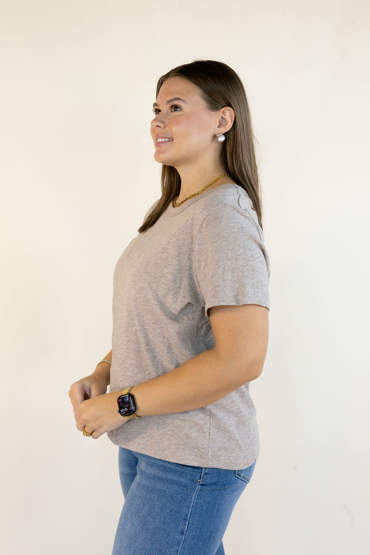 A woman wearing a light ash heather crewneck t-shirt and blue jeans, shown in profile against a plain off-white background.