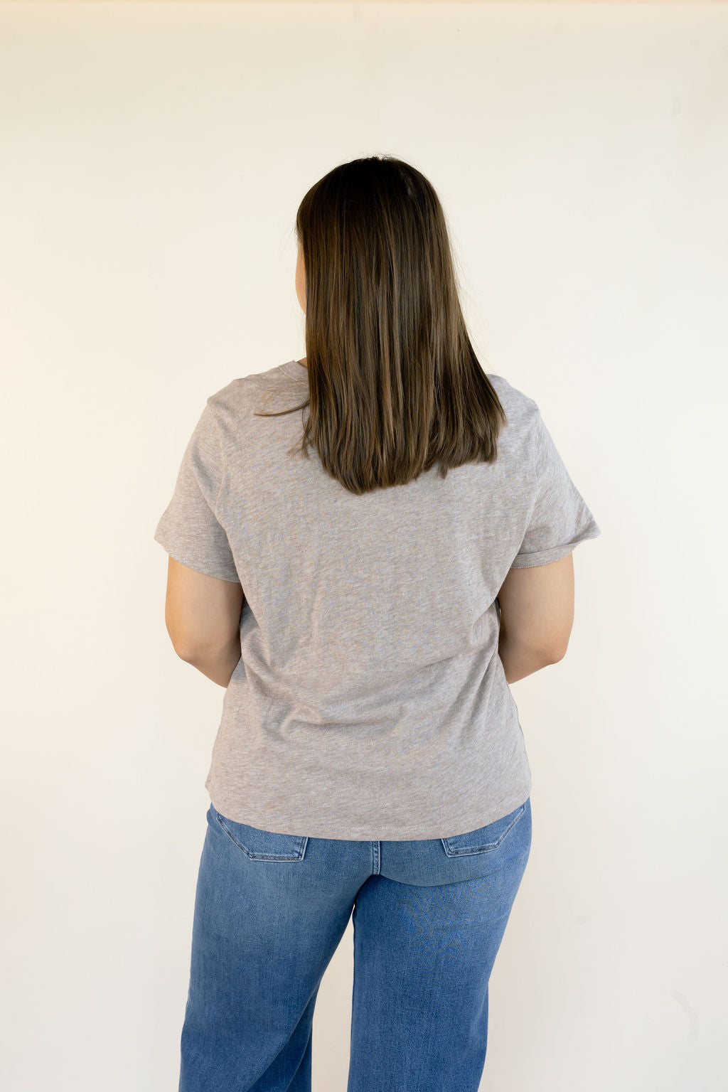 A woman viewed from behind wearing a light ash heather crewneck t-shirt and blue jeans, standing against a plain off-white wall.