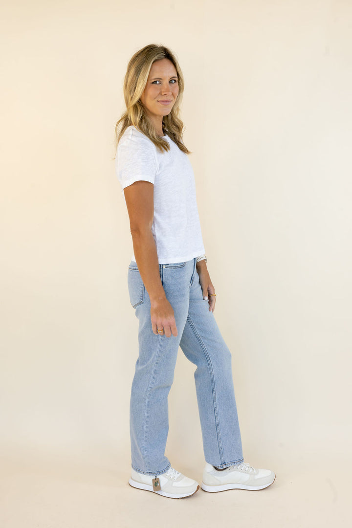 A woman modeling a white short-sleeve t-shirt and light-wash blue jeans, standing in profile against a solid beige studio background.