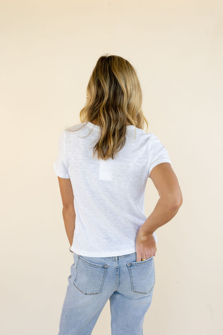 A woman with blonde-highlighted hair is shown from behind wearing a white short-sleeve t-shirt and light-wash blue jeans against a plain beige background.