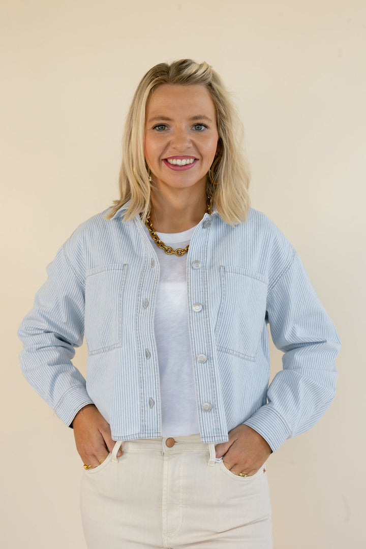 A smiling blonde woman wearing a light blue and white vertically striped cropped button-up jacket over a white t-shirt and white jeans, standing against a solid beige background.