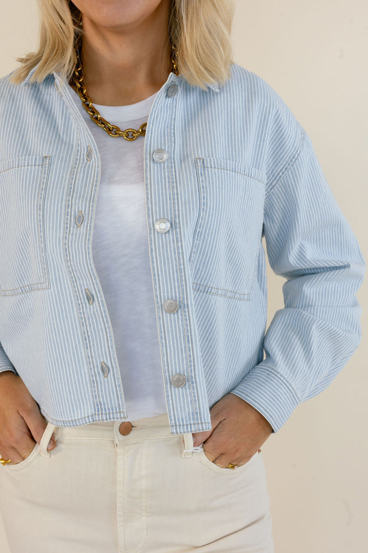 A person wearing a light blue and white vertically striped cropped denim jacket over a white t-shirt, paired with cream-colored jeans and a chunky gold chain necklace, against a plain beige background.