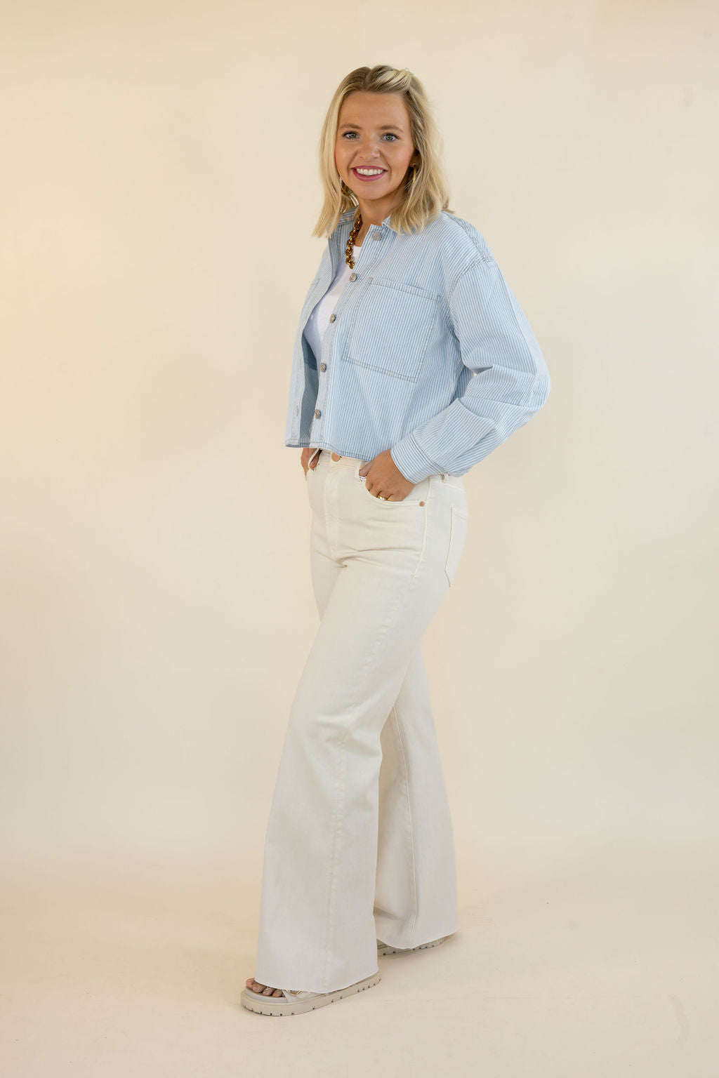 A woman modeling a light blue cropped denim  jacket and white wide-leg jeans against a plain beige studio background.