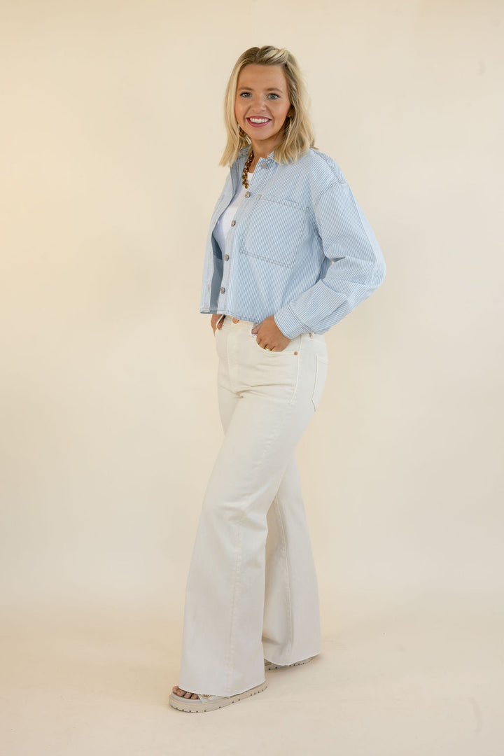 A woman modeling a light blue cropped denim  jacket and white wide-leg jeans against a plain beige studio background.