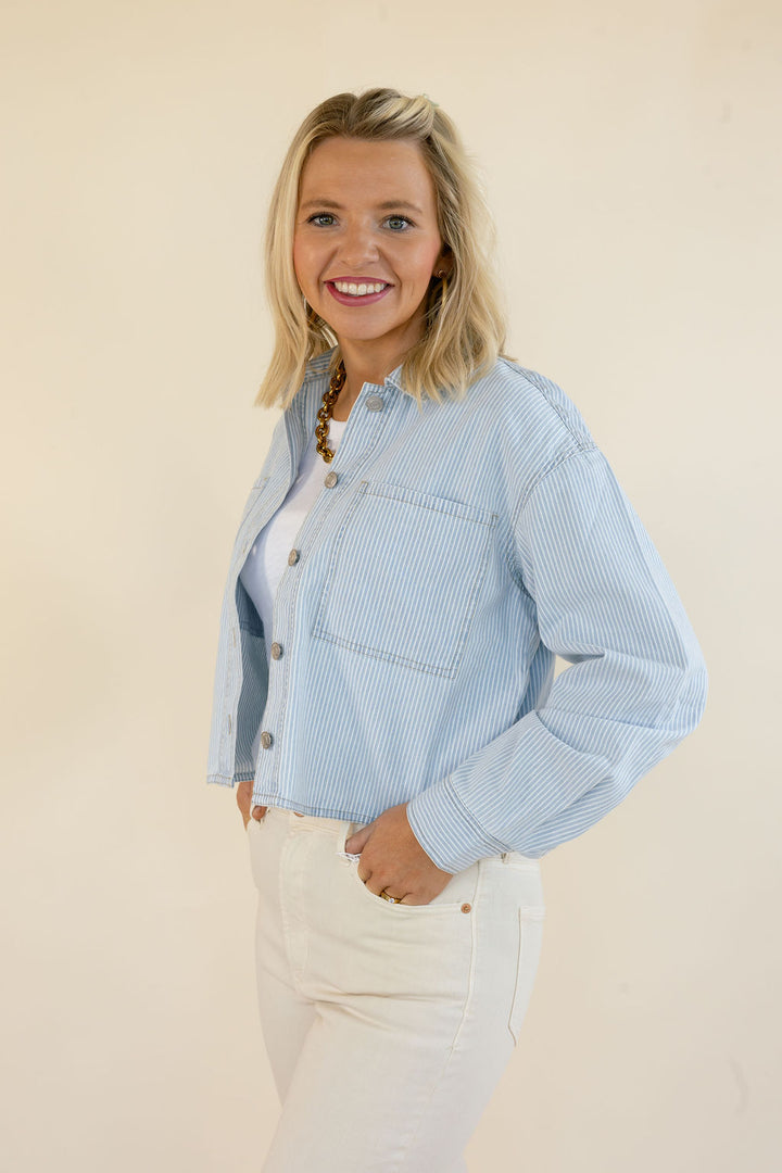 A smiling blonde woman wearing a light blue and white vertically striped cropped button-up jacket over a white top and white pants, standing against a plain beige studio background.