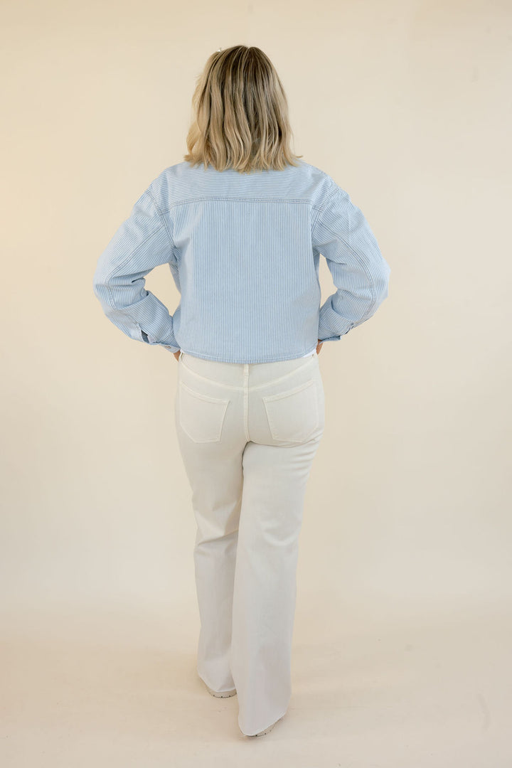 A woman viewed from behind wearing a light blue long-sleeve button-up jacket and off-white flared jeans, standing against a plain beige studio background.