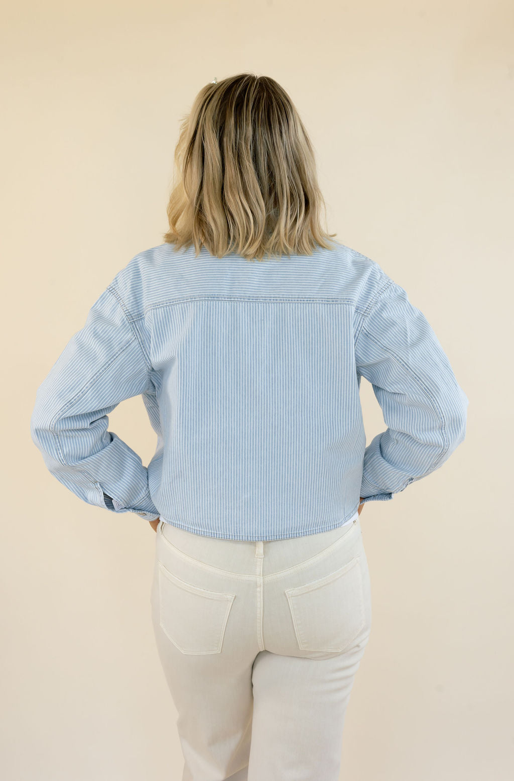 A woman viewed from behind wearing a light blue striped cropped jacket and cream-colored pants, standing against a solid beige background.
